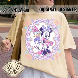May include: Cream-colored t-shirt featuring a Minnie Mouse and Daisy Duck graphic within a decorative frame. The design includes bows, hearts, and floral elements. The text "ORIGINAL DESIGNER" is visible. The shirt has a relaxed fit.