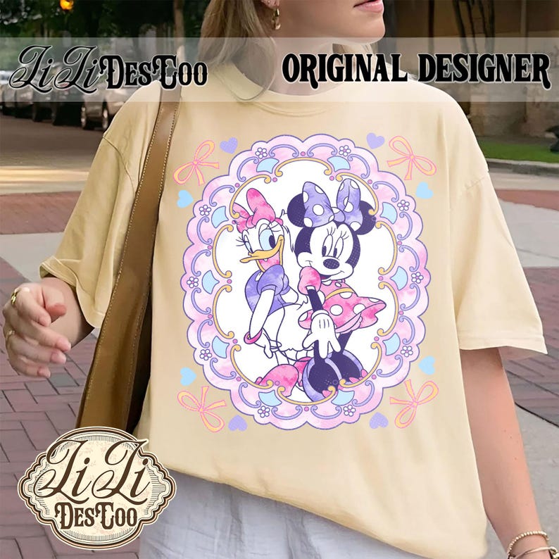May include: Cream-colored t-shirt featuring a Minnie Mouse and Daisy Duck graphic within a decorative frame. The design includes bows, hearts, and floral elements. The text "ORIGINAL DESIGNER" is visible. The shirt has a relaxed fit.