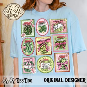 May include: Light blue t-shirt featuring a colorful design of pickle jars with pink bows and floral accents. The jars are labeled "Pickles" and "Dill Pickles". The shirt also includes the text "Li Li Des Too" and "Original Designer".