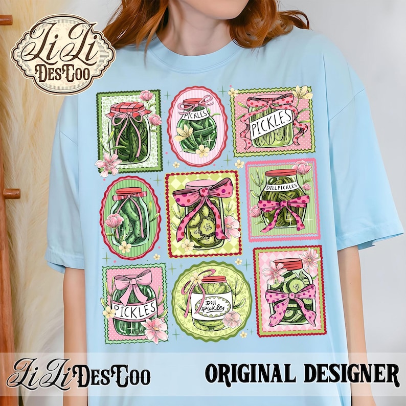 May include: Light blue t-shirt featuring a colorful design of pickle jars with pink bows and floral accents. The jars are labeled "Pickles" and "Dill Pickles". The shirt also includes the text "Li Li Des Too" and "Original Designer".