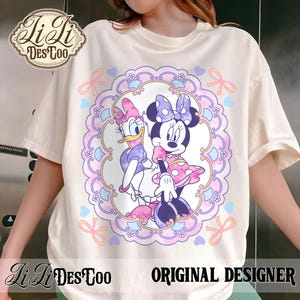 May include: Cream-colored t-shirt featuring a circular design with Minnie Mouse and Daisy Duck. The design is framed by a pink and purple border with bows and hearts. The shirt also has the text "LiLi DesToo ORIGINAL DESIGNER".