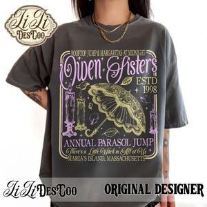 May include: A charcoal gray t-shirt featuring a graphic print with the text "Owen Sisters", "Rooftop Jump & Margaritas at Midnight", "Estd 1998", "Annual Parasol Jump", "There's a Little Witch in All of Us", and "Maria's Island, Massachusetts".