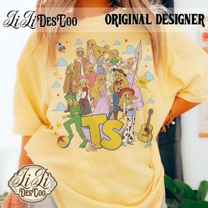 May include: A pale yellow t-shirt featuring a graphic print of multiple blonde characters in an anime style, including various outfits and accessories like a guitar, a sheriff badge, and a rocket. The large yellow letters TS are centered below.