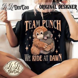 May include: Black t-shirt with the text "TEAM PUNCH" and the phrase "WE RIDE AT DAWN". The shirt features a graphic of two monkeys hugging, surrounded by star shapes. The brand name "Li Li Des Too" is also visible.