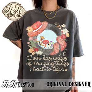 Puede incluir: Una camiseta gris carb&oacute;n con un estampado gr&aacute;fico de un marco de neum&aacute;tico que contiene personajes de dibujos animados, un sombrero de vaquero rojo y botas de vaquero rojas. El dise&ntilde;o incluye detalles florales y el texto Love has ways of bringing things back to life.
