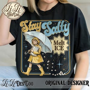 Pode incluir: Camiseta preta com um gr&aacute;fico que apresenta uma crian&ccedil;a com capa de chuva e botas amarelas segurando um guarda-chuva. O texto "Stay Salty Melt ICE" &eacute; exibido em uma fonte retr&ocirc;. O design inclui estrelas e gelo caindo.