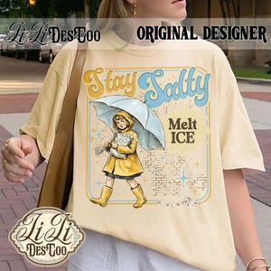 Pode incluir: Camiseta creme com um gr&aacute;fico apresentando uma menina de capa de chuva e botas amarelas segurando um guarda-chuva. O texto "Stay Salty Melt ICE" &eacute; exibido em uma fonte retr&ocirc;. A camisa tem um caimento folgado.