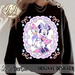 May include: Black sweatshirt featuring a Minnie Mouse and Daisy Duck graphic within a decorative oval frame. The design includes bows, hearts, and the text "LiLi DesToo ORIGINAL DESIGNER". The sweatshirt is a casual wear item.