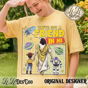 Puede incluir: Camiseta amarilla mostaza con el texto "YOU'VE GOT A FRIEND IN ME" y un gr&aacute;fico de Jes&uacute;s tomados de la mano con dos personajes de dibujos animados. El dise&ntilde;o incluye un cohete, planetas y estrellas. La camiseta tiene el logo "LiLi DesCoo" y el texto "ORIGINAL DESIGNER".