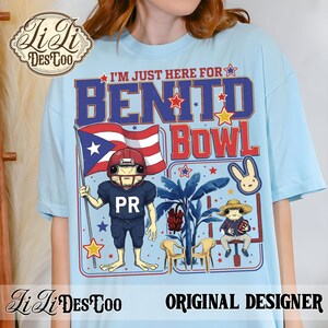 May include: Light blue t-shirt with a graphic featuring a frog in a football uniform holding a Puerto Rican flag. Text reads "I'M JUST HERE FOR BENITO BOWL". Includes a bunny and frog in a hat. "Li Li DesCoo" and "ORIGINAL DESIGNER" are also printed.