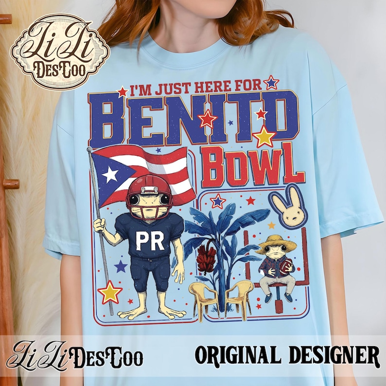 May include: Light blue t-shirt with a graphic featuring a frog in a football uniform holding a Puerto Rican flag. Text reads "I'M JUST HERE FOR BENITO BOWL". Includes a bunny and frog in a hat. "Li Li DesCoo" and "ORIGINAL DESIGNER" are also printed.