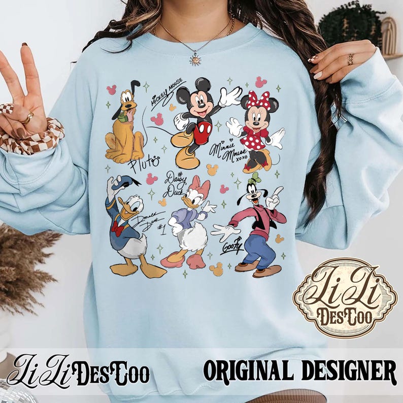 Puede incluir: Sudadera azul claro con un estampado colorido de personajes de Disney, incluyendo Mickey Mouse, Minnie Mouse, Pluto, Donald Duck, Daisy Duck y Goofy. Tambi&eacute;n se incluyen los nombres de los personajes. El texto "Original Designer" tambi&eacute;n es visible.