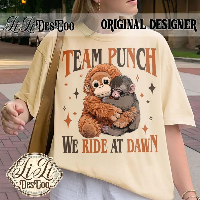 May include: Cream-colored t-shirt featuring a graphic of two hugging monkeys with the text "TEAM PUNCH" and "WE RIDE AT DAWN" in brown and black. The shirt also has star and diamond accents. The brand name "Li Li DesToo" is also visible.