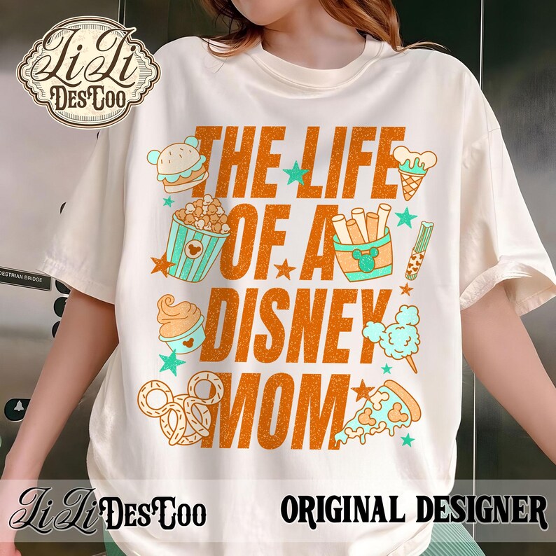 May include: Cream-colored t-shirt with the text "THE LIFE OF A DISNEY MOM" in orange, featuring food graphics like a burger, popcorn, and pizza. The shirt also has the designer's logo and the words "ORIGINAL DESIGNER".