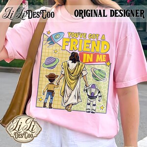 Puede incluir: Camiseta rosa claro con un dise&ntilde;o gr&aacute;fico que incluye el texto "YOU'VE GOT A FRIEND IN ME", un cohete, planetas y personajes de dibujos animados. La camiseta tambi&eacute;n tiene el texto "ORIGINAL DESIGNER" y la marca "Li Li DesCoo".