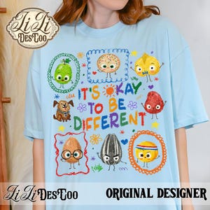 May include: Light blue t-shirt featuring the phrase "IT'S OKAY TO BE DIFFERENT" with colorful illustrations of various food items and a dog. The shirt has a relaxed fit and a crew neck.