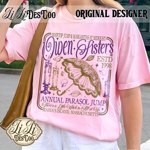 May include: A light pink t-shirt featuring a graphic print with the text "Owen Sisters", "Rooftop Jump & Margaritas at Midnight", "ESTD 1998", "Annual Parasol Jump", "There's a Little Witch in All of Us", and "Maria's Island, Massachusetts" with candles and a parasol.
