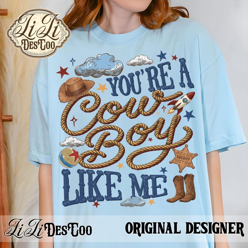 May include: A light blue t-shirt featuring a graphic print with the text "YOU'RE A COWBOY LIKE ME" in dark blue letters. The design includes a brown cowboy hat, a rocket, a sheriff badge, a ball, clouds, stars, and brown rope accents.
