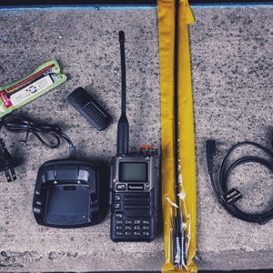 Könnte beinhalten: Ein schwarzes Funkgerät mit Antenne, Ladegerät und Zubehör. Das Funkgerät hat einen Bildschirm und nummerierte Tasten. Enthalten sind eine Ladestation, eine Ersatzantenne in einem gelben Etui und ein Kabel.