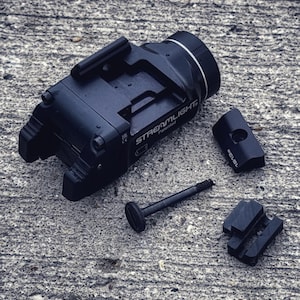 May include: A black Streamlight TLR-1 HL weapon light with a black polymer body and a silver bezel. The light is attached to a black rail mount with a black screw. The light is on a grey concrete surface.