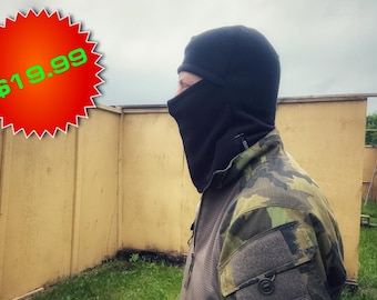 May include: A person wearing a black balaclava and camouflage jacket. The balaclava covers the head and neck, with only the face visible. The jacket has a green, brown, and tan camouflage pattern. The background shows a wooden structure.