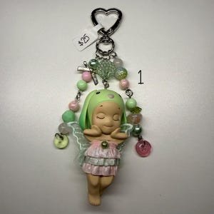 Puede incluir: Un llavero caprichoso con una peque&ntilde;a figura de hada con un sombrero de conejito verde y un vestido rosa. El llavero incluye un cierre en forma de coraz&oacute;n, cuentas decorativas en colores pastel y una peque&ntilde;a etiqueta.