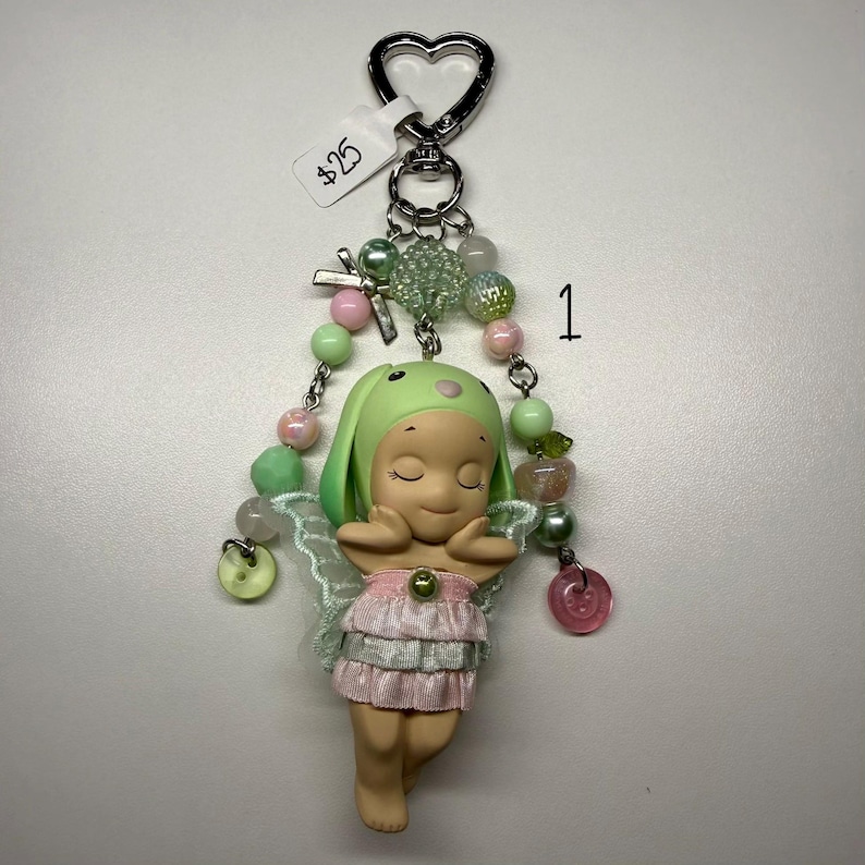 Puede incluir: Un llavero caprichoso con una peque&ntilde;a figura de hada con un sombrero de conejito verde y un vestido rosa. El llavero incluye un cierre en forma de coraz&oacute;n, cuentas decorativas en colores pastel y una peque&ntilde;a etiqueta.