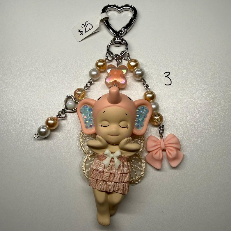 Puede incluir: Un llavero caprichoso con una figura de hada elefante rosa con los ojos cerrados, una falda con volantes y un lazo. El llavero incluye un cierre en forma de coraz&oacute;n, detalles de perlas y cuentas, y un dije de lazo rosa.