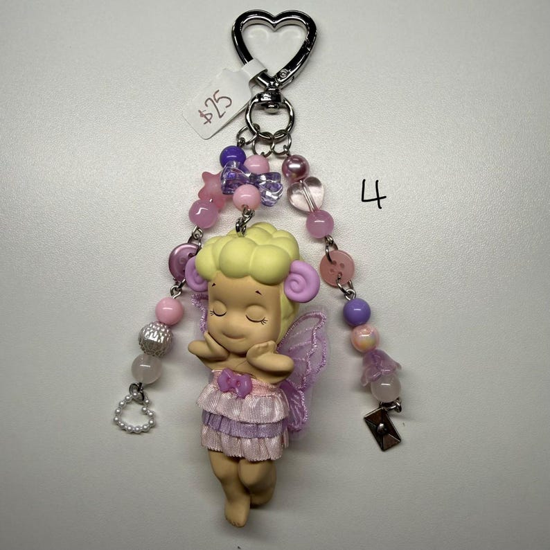 Puede incluir: Un llavero caprichoso con una figura de hada con los ojos cerrados, cabello amarillo claro, alas moradas y un vestido rosa con volantes. El llavero incluye un cierre en forma de coraz&oacute;n y detalles de cuentas en tonos rosa, morado y plateado.