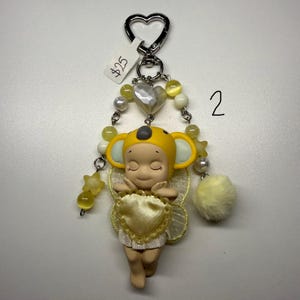 Puede incluir: Un llavero caprichoso con una peque&ntilde;a figura con un sombrero de koala amarillo y alas en forma de coraz&oacute;n. El llavero incluye un cierre plateado en forma de coraz&oacute;n, cuentas amarillas y un pomp&oacute;n amarillo esponjoso. El n&uacute;mero "2" est&aacute; escrito en el fondo.