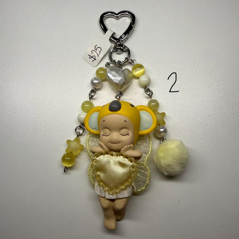 Puede incluir: Un llavero caprichoso con una peque&ntilde;a figura con un sombrero de koala amarillo y alas en forma de coraz&oacute;n. El llavero incluye un cierre plateado en forma de coraz&oacute;n, cuentas amarillas y un pomp&oacute;n amarillo esponjoso. El n&uacute;mero "2" est&aacute; escrito en el fondo.