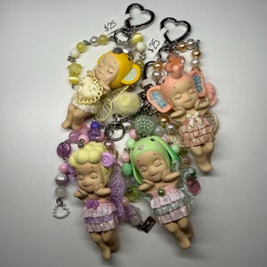 Puede incluir: Cuatro llaveros con figuras de beb&eacute;s durmientes con alas y sombreros. Cada llavero tiene un esquema de color diferente, incluyendo amarillo, rosa, verde y morado. Los llaveros est&aacute;n adornados con cuentas y dijes, y tienen cierres en forma de coraz&oacute;n.