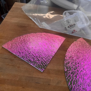 May include: Two iridescent pink glass pieces with a textured surface, shaped like quarter circles, are displayed on a wooden surface. A clear plastic bag and a label are in the background.