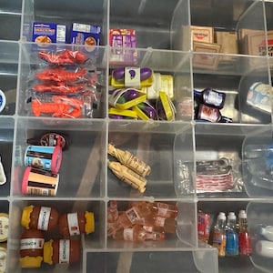 May include: A clear plastic organizer filled with miniature food items. Includes boxes of pretzels, packages of lobster, containers of whipped cream, and bottles of water. Other items include miniature pizza boxes and bottles of kombucha.