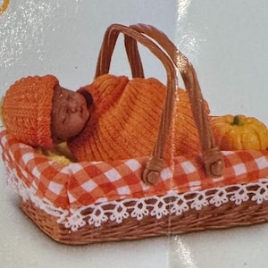 May include: A miniature figurine of a baby sleeping inside a woven basket. The baby wears an orange knit hat and is wrapped in an orange blanket. The basket features an orange and white gingham fabric liner with white lace trim and a small pumpkin.