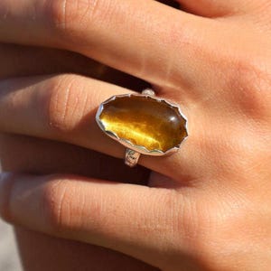 May include: A silver ring featuring a large, oval-shaped, translucent golden-brown gemstone with a smooth, domed surface. The stone is set within a scalloped, serrated silver bezel, and the ring band has a textured, hammered finish.