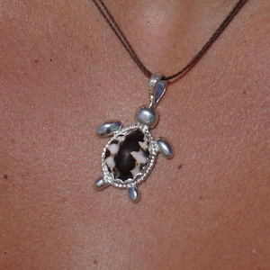 May include: A silver turtle pendant necklace featuring a dark brown and white patterned shell inlay. The turtle charm is suspended from a thin, dark brown cord. The metal frame of the turtle is polished silver, highlighting the four flippers and head.