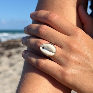 Pode incluir: Um anel prateado com uma concha branca e bege. O anel é usado numa mão, com uma praia e o mar ao fundo. O design do anel realça a beleza natural da concha.