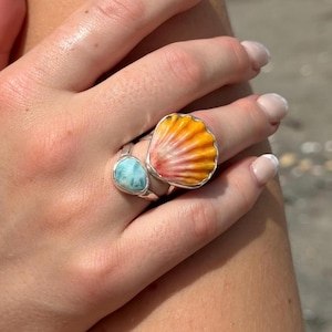 May include: A silver ring featuring two distinct stones: a small, teardrop-shaped light blue stone with white marbling and a larger, fan-shaped seashell with a gradient of yellow, orange, and pink hues, both set in polished silver metal bands.