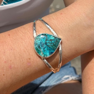 Puede incluir: Una pulsera tipo brazalete con una piedra turquesa en forma de lágrima con vetas oscuras, engastada en un marco de metal plateado pulido. La banda consta de dos alambres de metal plateado martillado que se unen en el engaste central.
