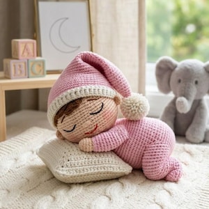May include: A handmade crochet doll in a sleeping pose, wearing a pink outfit and hat with a white trim and pom-pom. The doll is lying on a cream-colored pillow and blanket. A gray stuffed elephant is in the background.