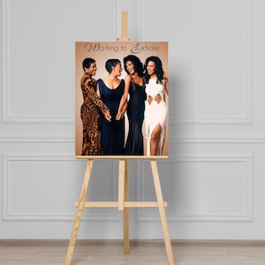 Puede incluir: Un caballete de madera exhibe una impresión con cuatro mujeres en elegantes vestidos. Las mujeres se toman de la mano y sonríen. La impresión tiene las palabras "Waiting to Exhale" en una fuente decorativa.