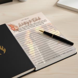 Może przedstawiać: Czarny notatnik z liniowanymi stronami i tekstem "Writing to Exhale" i "Let it Burn!". Czarny i złoty długopis spoczywa na otwartych stronach. Okładka notatnika jest czarna ze złotym emblematem.
