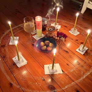 Op de afbeelding: Een rituele scène op een houten vloer, met een cirkel getekend met krijt, brandende kaarsen in glazen houders, een wijnglas, een kom met vijgen en een grote kaars. Er stijgt wierookrook op en er zijn gedroogde bloemen aanwezig.