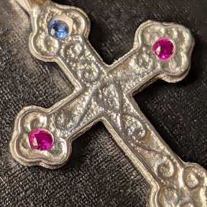 May include: A silver-toned metal cross pendant featuring an intricate embossed scrollwork pattern across its surface. The cross is accented with three small, round, faceted gemstones: one light blue stone at the top and two vibrant pink stones.