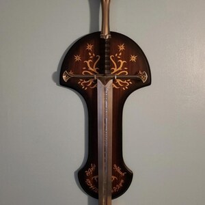May include: A silver sword with a black handle is mounted on a dark brown wooden wall plaque. The plaque features a gold tree design and the inscription "Ash Nazg Durbatul&ucirc;k, Ash Nazg Gimbatul, Ash Nazg Thrakul&ucirc;k, Agh Burzum-ishi krimpatul."
