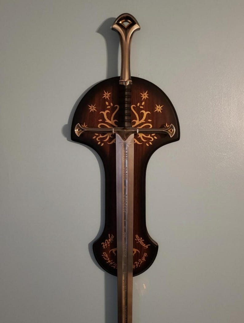 May include: A silver sword with a black handle is mounted on a dark brown wooden wall plaque. The plaque features a gold tree design and the inscription "Ash Nazg Durbatul&ucirc;k, Ash Nazg Gimbatul, Ash Nazg Thrakul&ucirc;k, Agh Burzum-ishi krimpatul."