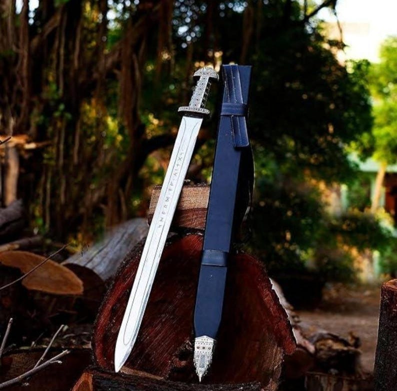 May include: A silver Viking sword with a black leather scabbard. The sword has runes engraved on the blade and a silver pommel. The scabbard has a silver tip and a black leather strap.