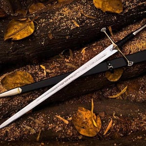 May include: A silver sword with a black leather scabbard and a gold-colored hilt. The sword is lying on a bed of brown wood chips and leaves.