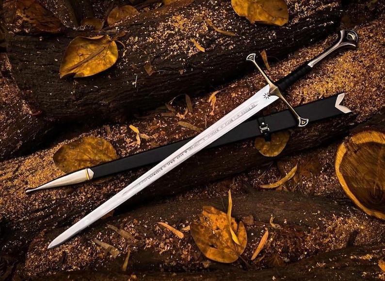 May include: A silver sword with a black leather scabbard and a gold-colored hilt. The sword is lying on a bed of brown wood chips and leaves.